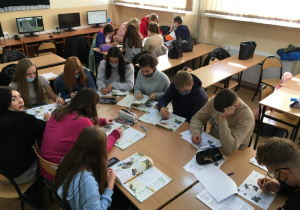 Uczniowie siedzą w grupach przy ławkach i rozwiązują zadania językowe w podręcznikach podczas kursu językowego w pracowni nr 2