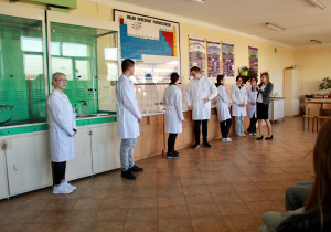 Uczniowie ubrani w białe fartuchy ochronne stoją przy stanowiskach chemicznych i demonstrują eksperymenty w pracowni 201 w budynku ZSP 8